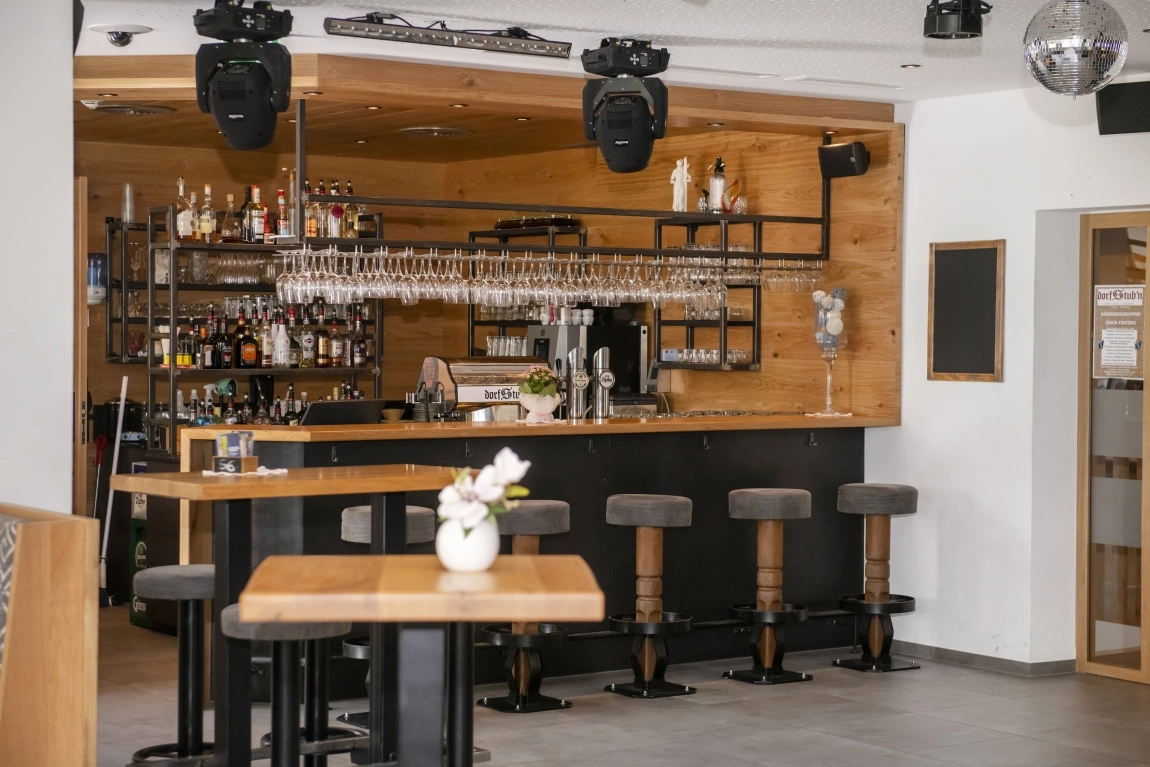Barbereich der dorfStub’n bei Tageslicht – moderne Holz- und Metallgestaltung, mit Barhockern, hoher Tischgarnitur und gut sortierter Getränkeauswahl.