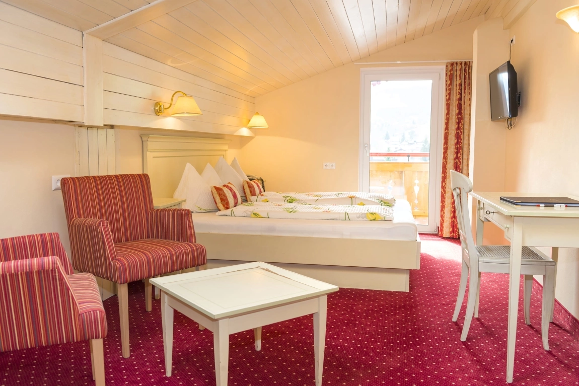 Helles Zimmer mit Dachschräge, Doppelbett, zwei rot gestreiften Sesseln, Tisch, Schreibtisch, Balkonzugang und Fernseher.