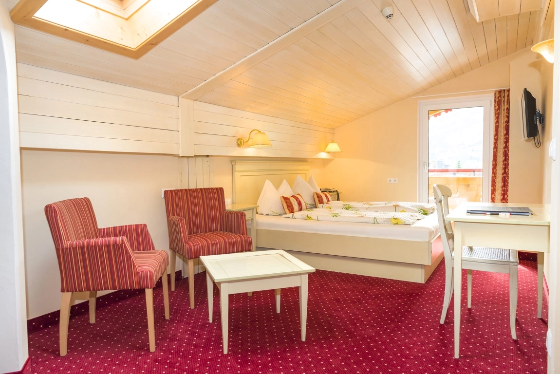 Dachzimmer mit rotem Teppich, zwei rotgestreiften Sesseln, hellem Tisch, Doppelbett und Zugang zum Balkon.