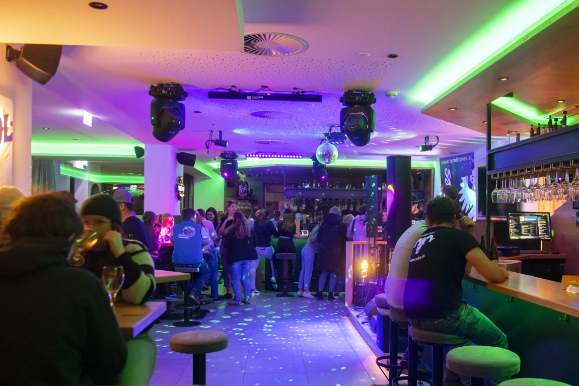 Stimmungsvolle Bar ‚dorfStub’n‘ mit grün-violetter Beleuchtung, Gästen an Tischen, Theke und auf der Tanzfläche.
