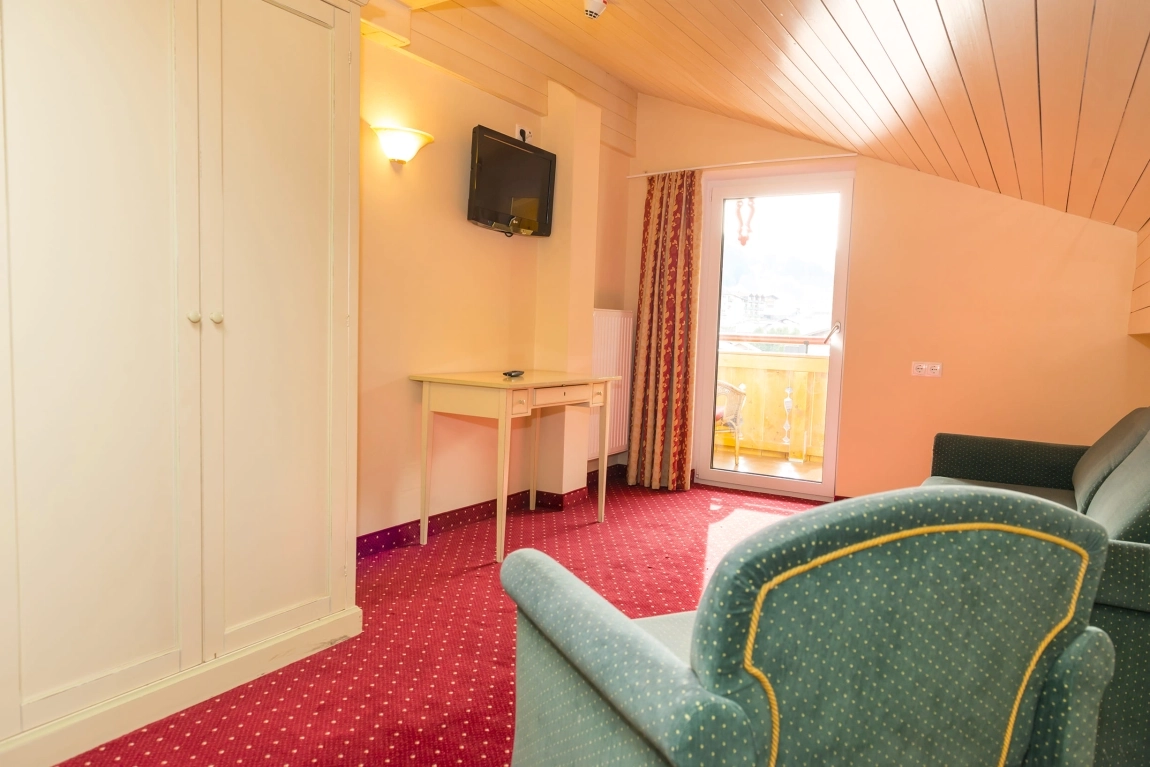 Gemütlicher Raum mit Dachschräge, grünem Sofa, TV, hellem Schreibtisch, Kleiderschrank und Zugang zum Balkon.