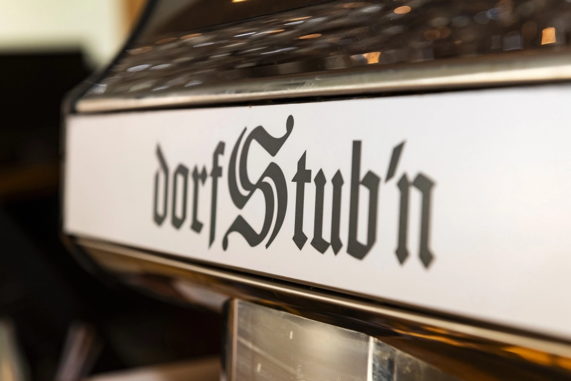 „Nahaufnahme des Schriftzugs ‚dorfStub’n‘ in traditioneller Schriftart auf hellem Hintergrund – gemütliches, ländliches Ambiente.