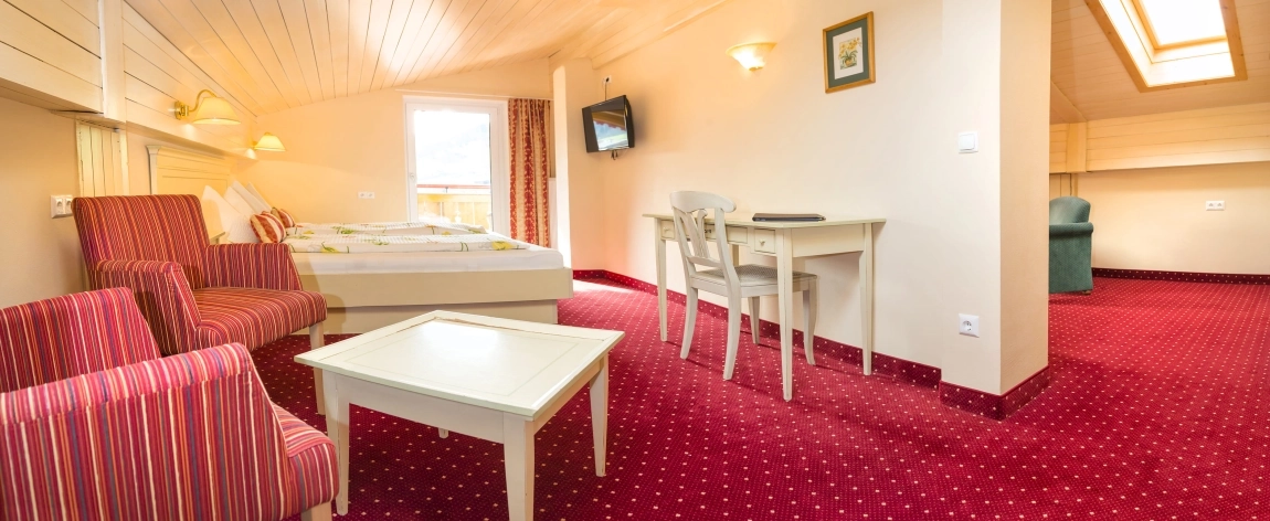 Dachzimmer mit rotem Teppich, Doppelbett, Esstisch, zwei Stühlen, TV an der Wand und Sofa im Nebenbereich.