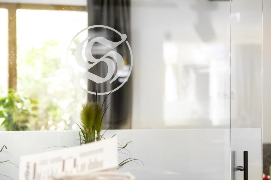 Glaswand mit stilisiertem „S“-Logo, dahinter Vorhänge, Pflanzen und Licht aus einem Fenster, alles in heller Umgebung.