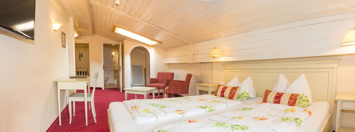 Dachzimmer mit Doppelbett, rotem Teppich, Tisch und Stühlen, helle Holzdecke und gemütlicher Sitzbereich.