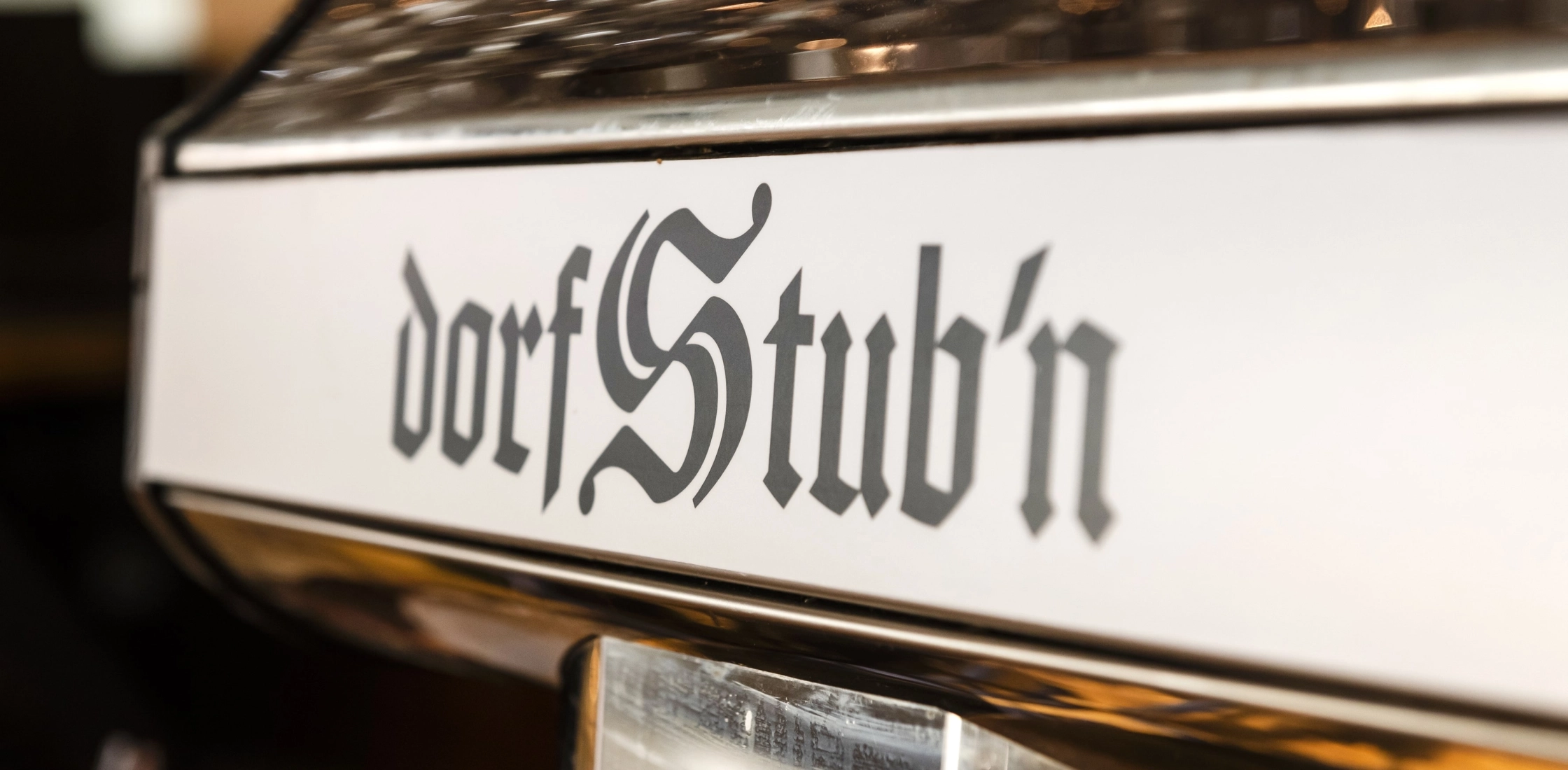 Nahaufnahme einer metallischen Fläche mit dem Schriftzug ‚dorfStub’n‘ in dekorativer Schrift, vermutlich Kaffeemaschine.