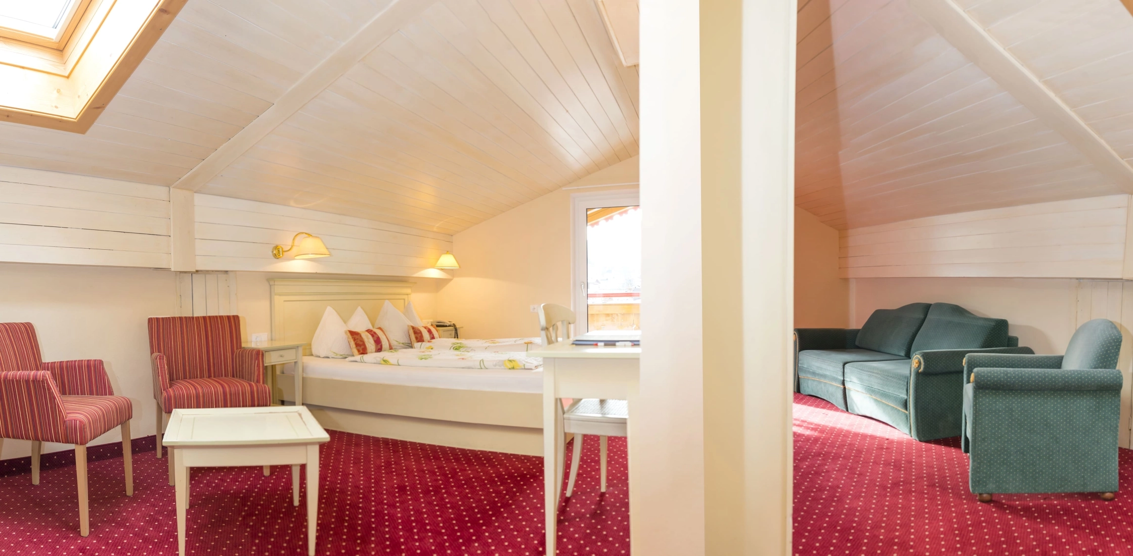 Dachzimmer mit rotem Teppich, Dachfenstern, Doppelbett, Sitzecke mit Sesseln und grünem Sofa im rechten Bereich.