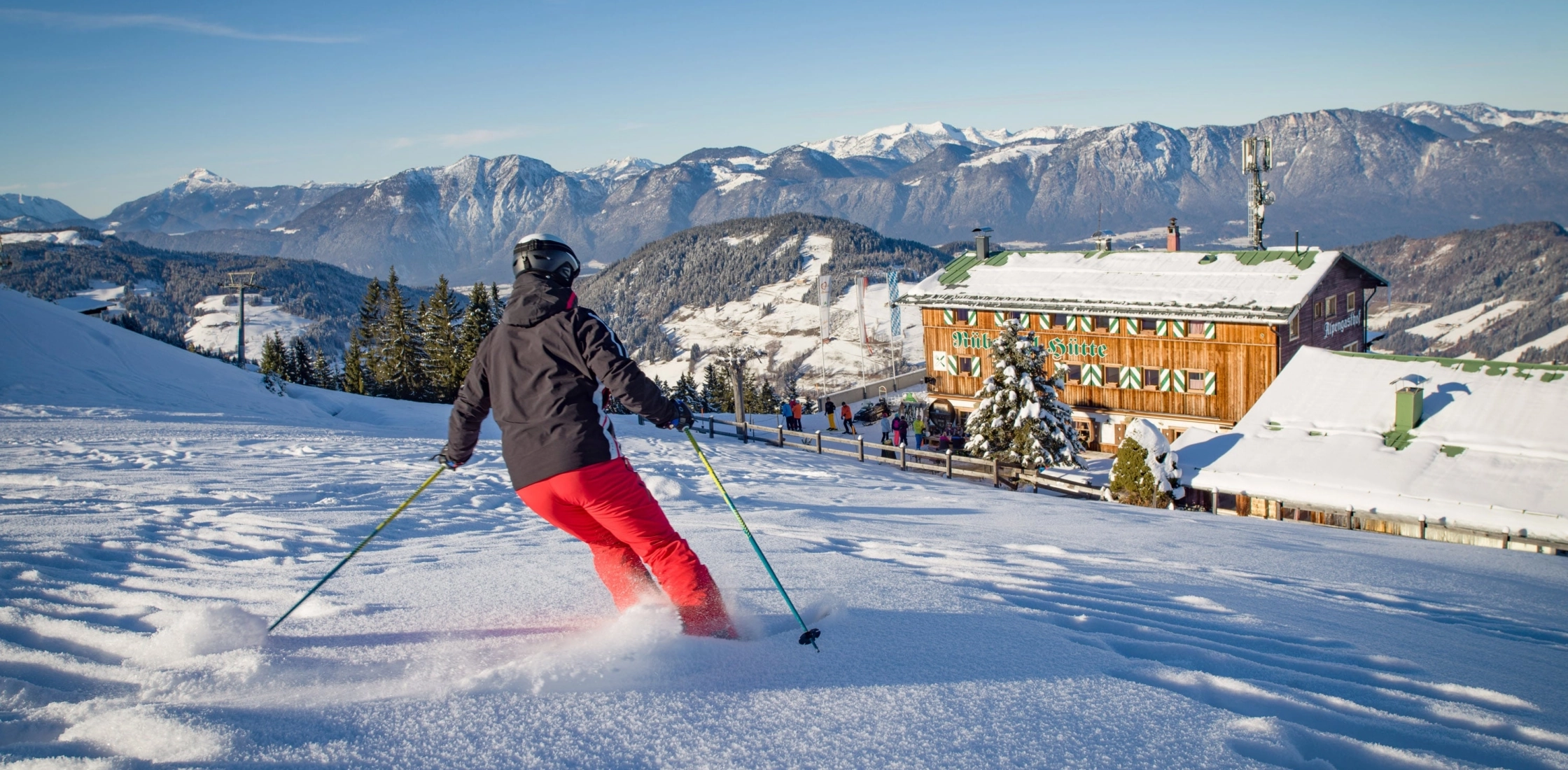 Ein Skifahrer in schwarzer Jacke und roten Hosen fährt auf einer verschneiten Piste, im Hintergrund ein gemütliches Berghotel und schneebedeckte Berge.