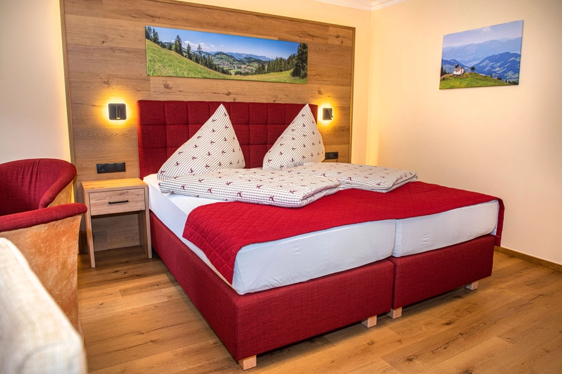 Gemütliches Doppelbett mit rotem Rahmen, gemustertem Bettzeug, Holzboden, Wandlampen und Landschaftsbildern an der Wand.