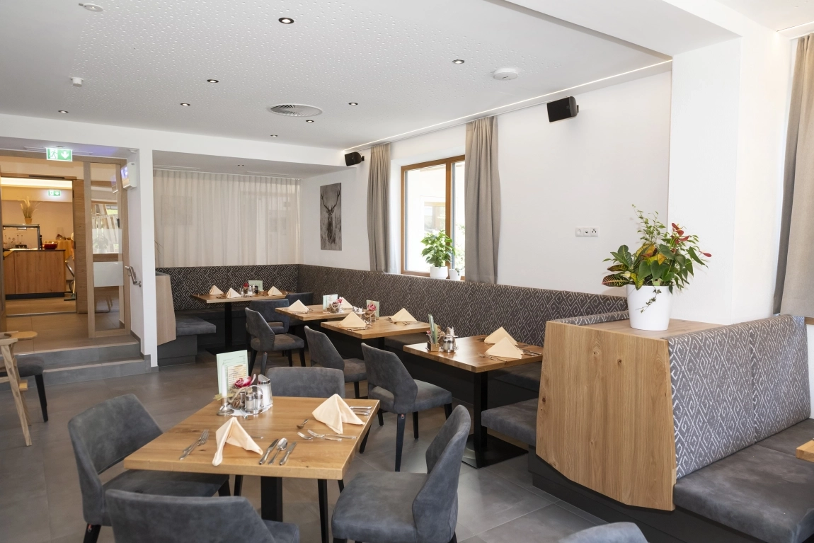 Gastraum mit Holztischen, gepolsterten Stühlen, langen Sitzbänken, Pflanzen und gut zugänglichem Durchgang.
