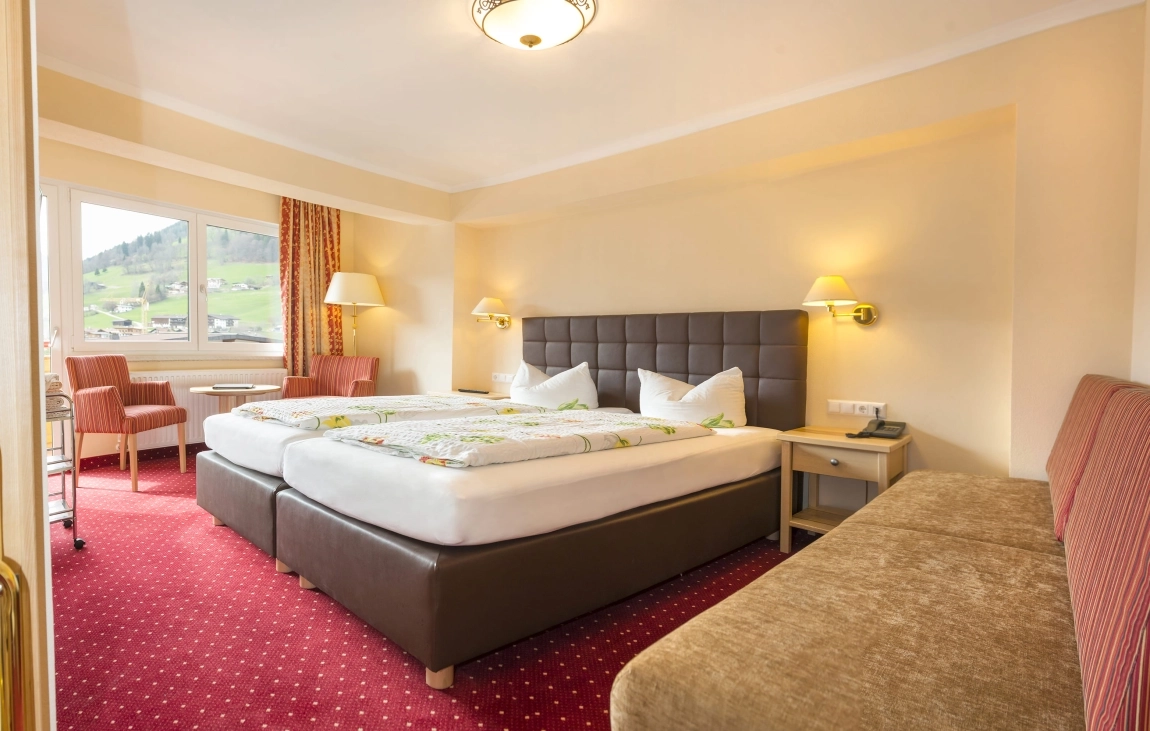 Hotelzimmer mit kontrastreichem Doppelbett (dunkles Kopfteil, helle Bettwäsche), rotem Teppich, Sofa, hellem Tageslicht und klaren Linien.