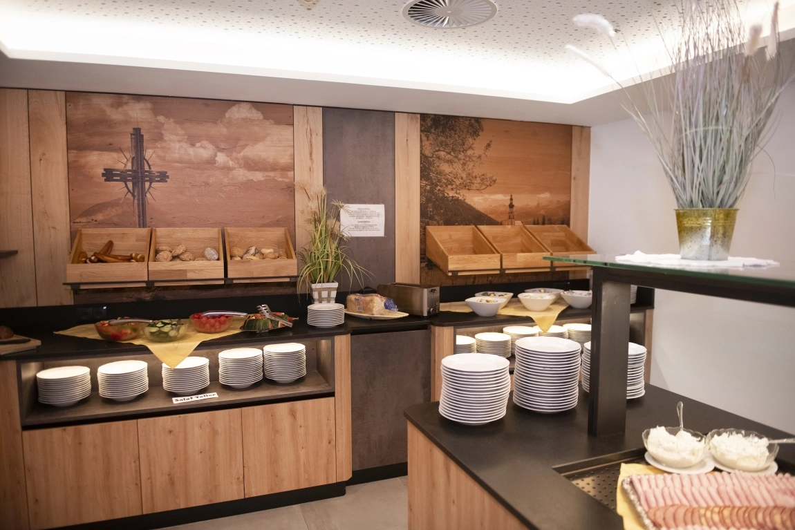 Frühstücksbuffet im Hotel mit Brotkörben, Müslibar, Getränken, Tellern, Besteck und Wandbild mit Kreuzmotiv.