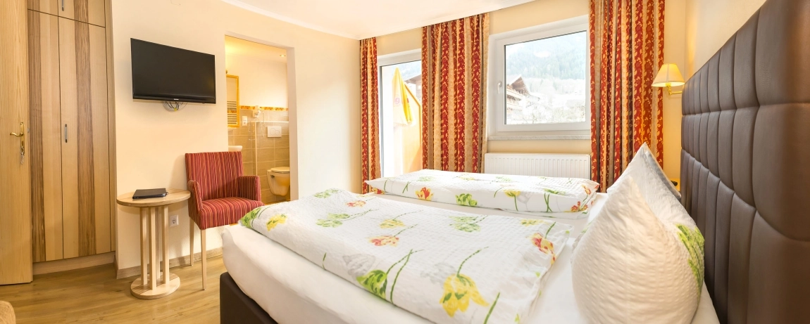 ChatGPT said: Helles Hotelzimmer mit Doppelbett, floraler Bettwäsche, zwei Fenstern, TV, Schrank und Zugang zum Bad.