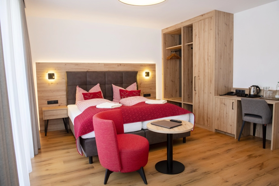 Modernes Zimmer mit Doppelbett, rotem Sessel, Tisch, Schreibtisch, offenem Kleiderschrank und Wasserkocher auf Holzfußboden.