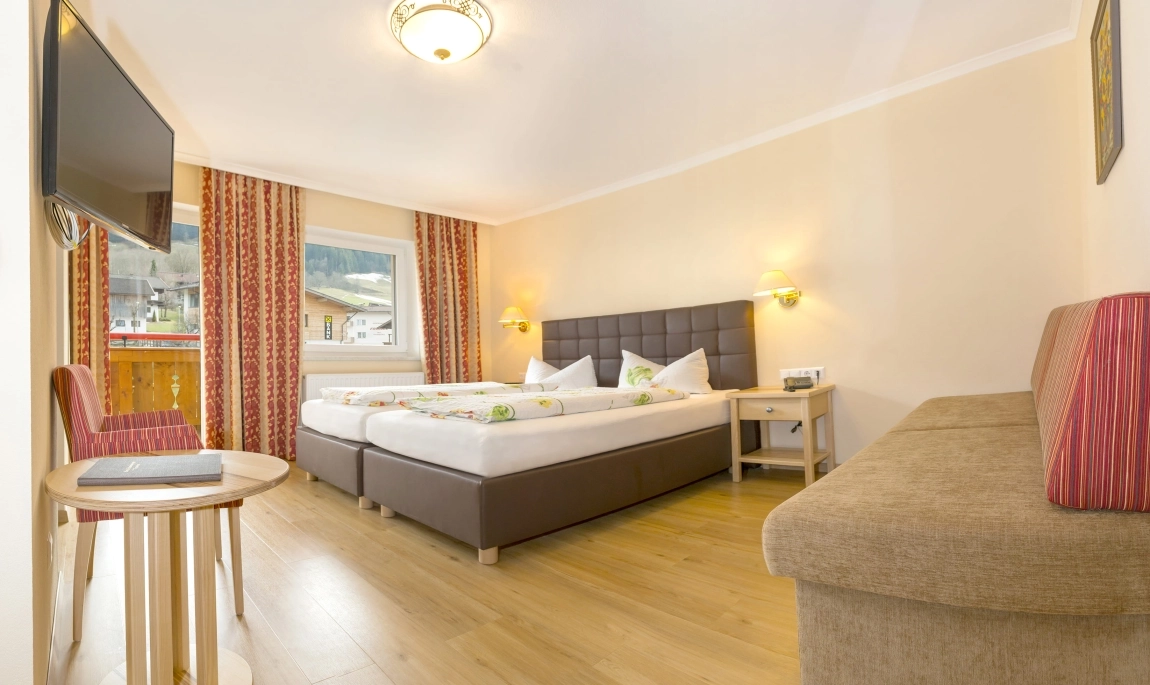 Helles Hotelzimmer mit Doppelbett, Couch, Flachbild-TV, Vorhängen in Rot-Gold und Blick auf die Berge durch das Fenster.