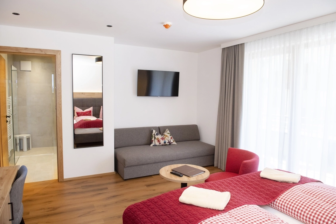 Modernes, helles Zimmer mit bodentiefem Fenster, kontrastreichem Bett, Sofa, TV und barrierefreier Dusche im Hintergrund.