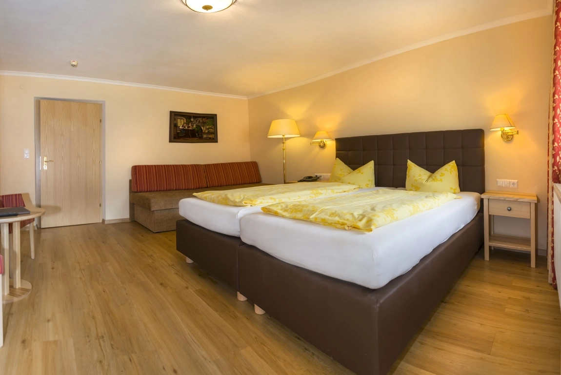 Geräumiges Hotelzimmer mit Doppelbett, gelber Bettwäsche, Sofa, Holzboden und warmem Licht an den Wänden.