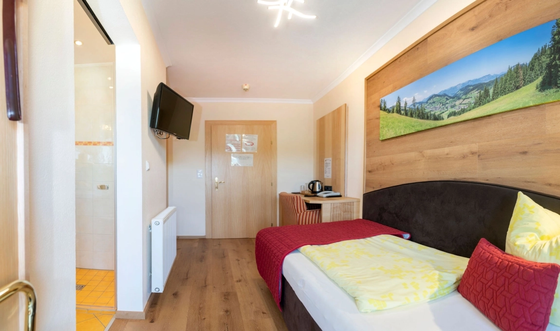 Helles Einzelzimmer mit Bett, Schreibtisch, TV und Badzugang. Warme Farben, klar strukturiert und barrierearm eingerichtet.