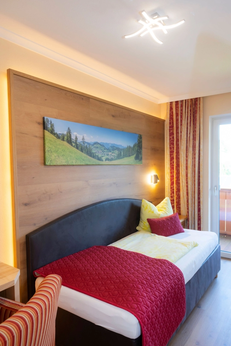 Einzelbett mit roter Decke, gelbem Kissen, hellem Holz, Naturbild an der Wand und Tür zum Balkon mit Tageslicht.
