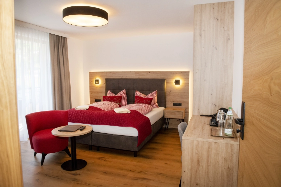Helles Zimmer mit Doppelbett, roter Sitzecke, Holzschreibtisch mit Wasserkocher, Vorhängen und Deckenleuchte auf Holzfußboden.