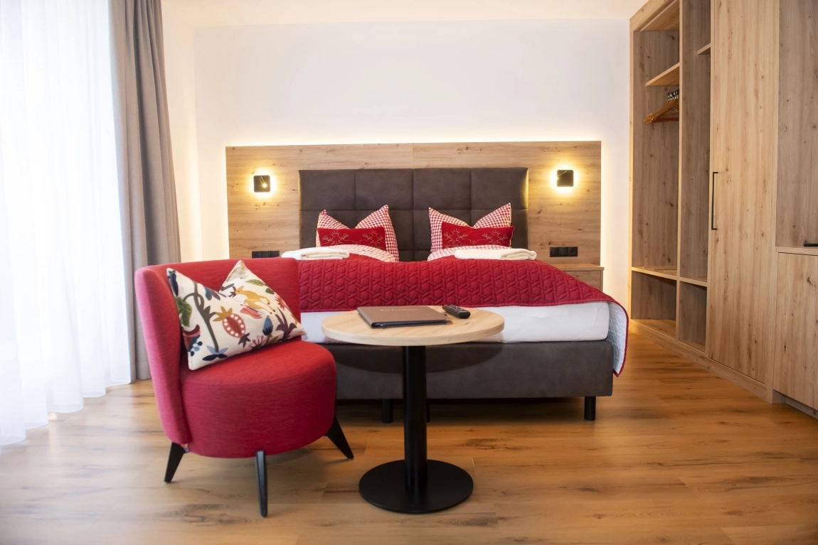 Helles Zimmer mit Doppelbett, rotem Sessel, kleinem Tisch, Holzschrank und Nachttischlampen an der Wand.