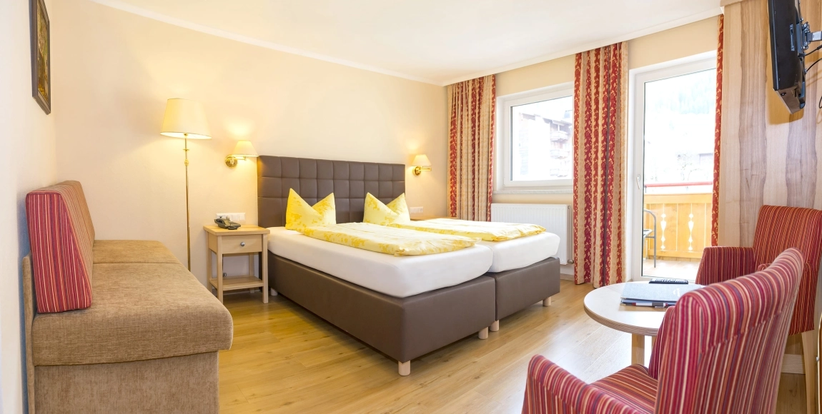 Helles Hotelzimmer mit Doppelbett, Couch, Sesseln, Tisch, Vorhängen in Rot-Gold und Zugang zum Balkon mit Ausblick.