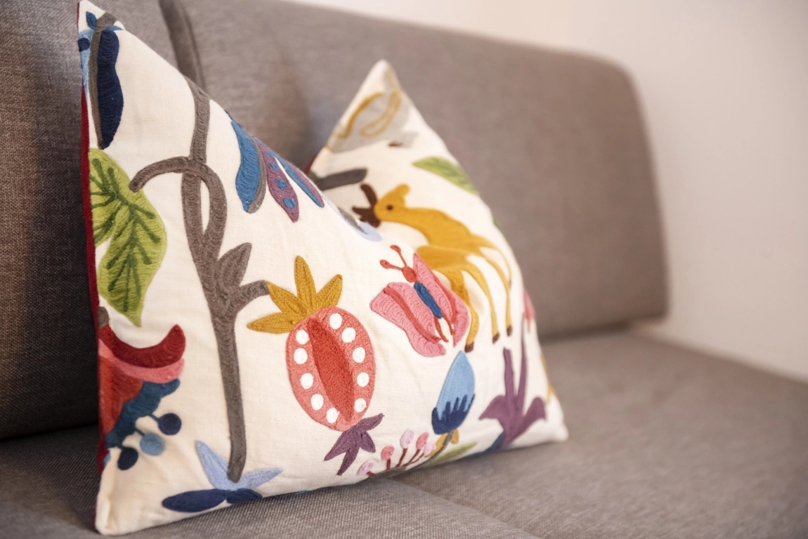 Dekokissen mit buntem, kontraststarkem Blumen- und Tiermuster liegt auf grauem Sofa mit glatter Struktur.