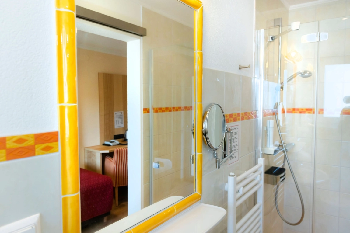 Helles Bad mit Dusche, gelb umrahmtem Spiegel, Handtuchheizung, Duschkopf und Blick ins angrenzende Zimmer.