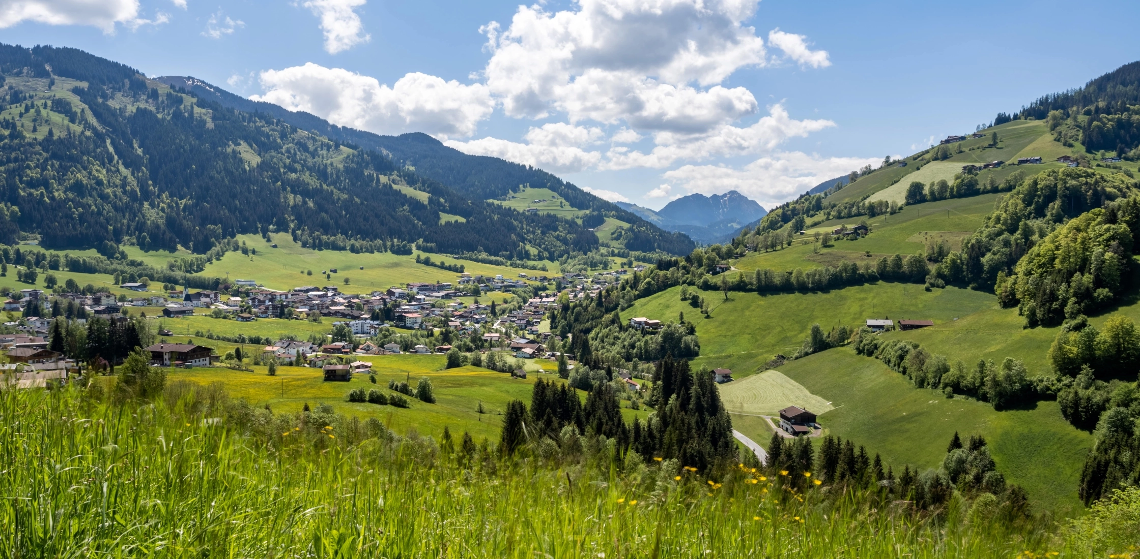 Ein grünes Tal mit einer kleinen Ortschaft, umgeben von Hügeln und Bergen, unter einem sonnigen, wolkigen Himmel.