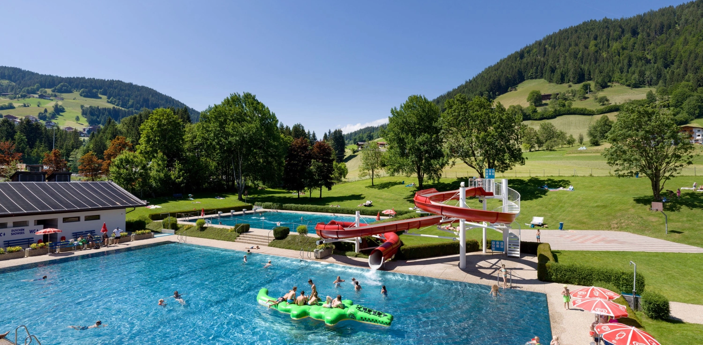 Ein Freibad mit Rutschen und Schwimmern, umgeben von grünen Hügeln und Bäumen bei strahlendem Sonnenschein.