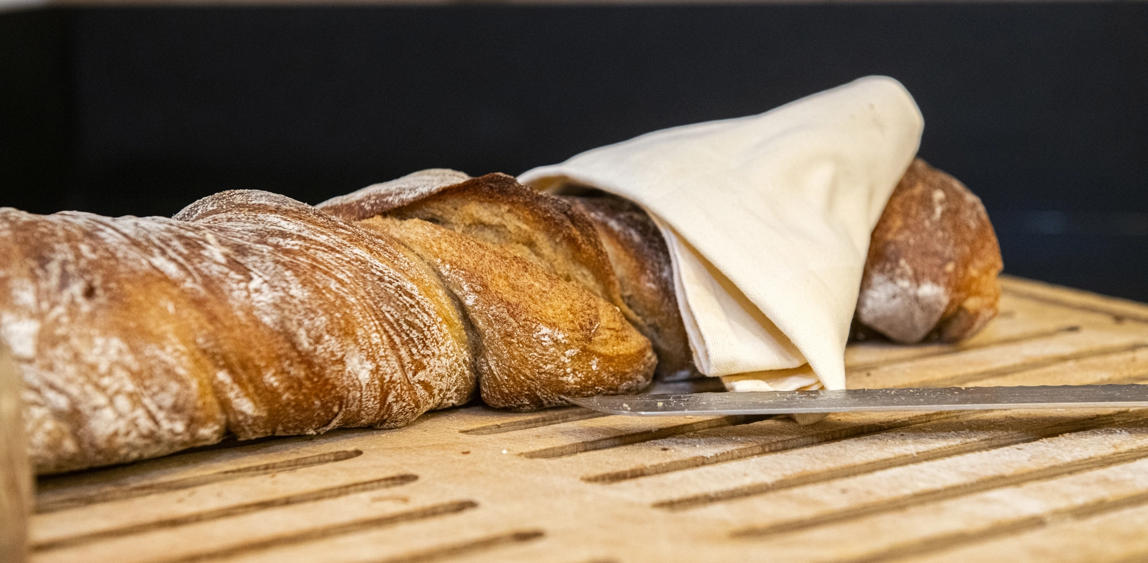Frisches Brot auf Holzbrett mit Messer und Serviette, bereit zur Selbstbedienung am Frühstücksbuffet.