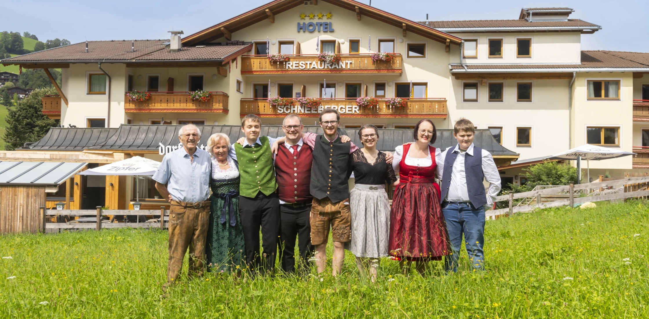 Das Bild zeigt die Familie, die das Hotel Schneeberger führt. Sie trägt traditionelle Trachten und steht vor dem Hotelgebäude auf einer grünen Wiese.