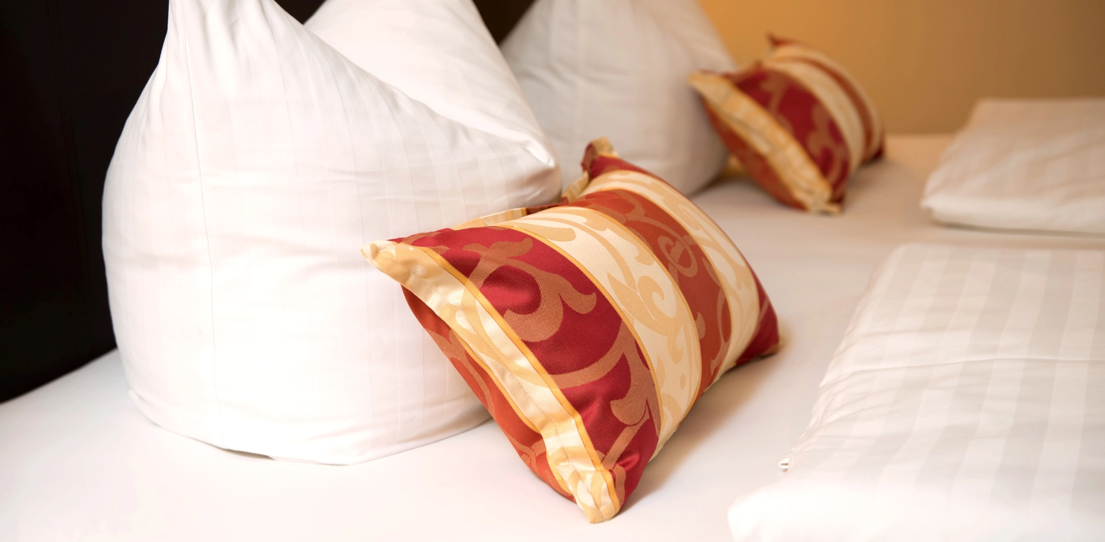 Nahaufnahme von weißem Bett mit Zierkissen in Rot-Gold und drei weißen Kopfkissen vor hellem Hintergrund.