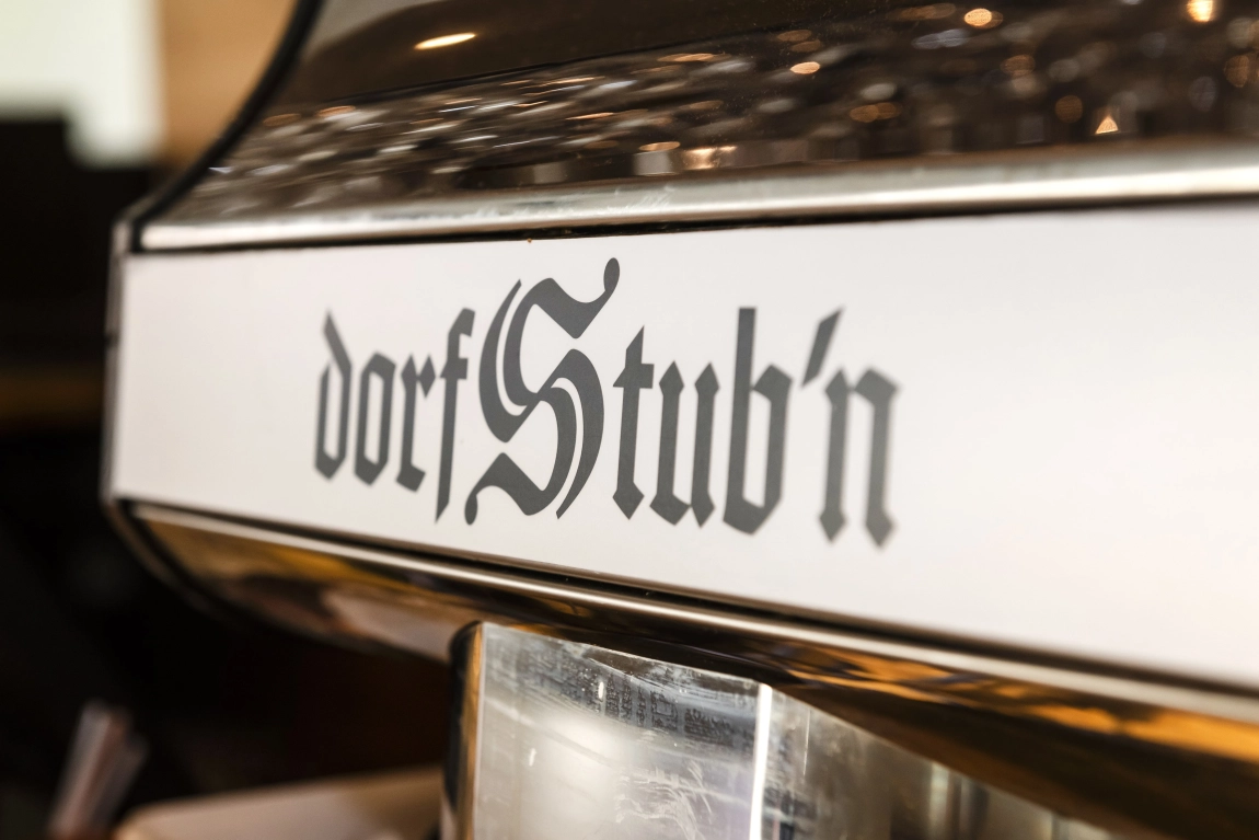 Nahaufnahme einer Metallfläche mit der Aufschrift ‚dorfStub’n‘ in altdeutscher Schrift, Teil einer Kaffeemaschine.