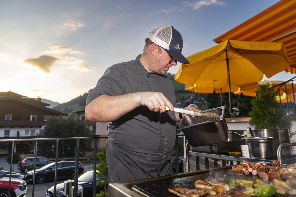 Hotelchef grillt auf der Terrasse bei Sonnenuntergang, mit Topf in der Hand, Sonnenschirm und Häuser im Hintergrund.