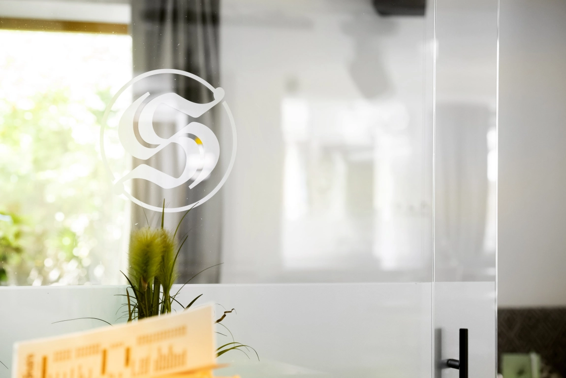 Glaswand mit großem S-Logo, niedriger Pflanze davor und Blick auf einen hellen, modern eingerichteten Innenraum