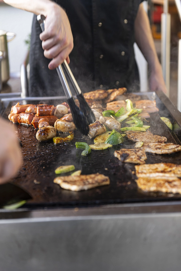 Person grillt Fleisch, Würstchen und Gemüse auf großer Grillplatte, dampfend und frisch zubereitet.
