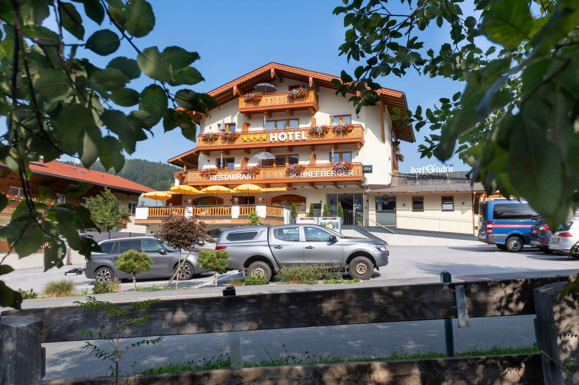 Hotel Schneeberger mit Balkonen, Sonnenterrasse, Parkplatz mit Autos und Blick durch Laub im Vordergrund.