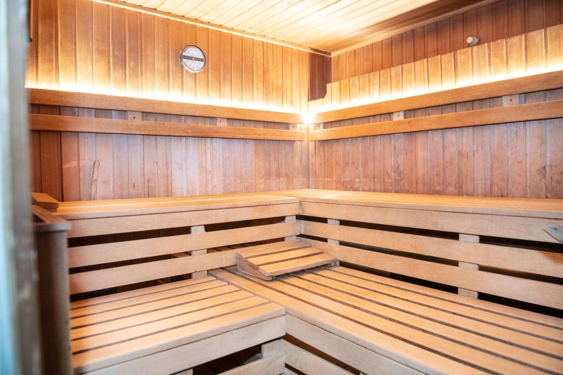 Helle Sauna aus Holz mit breiten Sitzflächen, Rückenlehnen, indirektem Licht und einem Thermometer an der Wand.