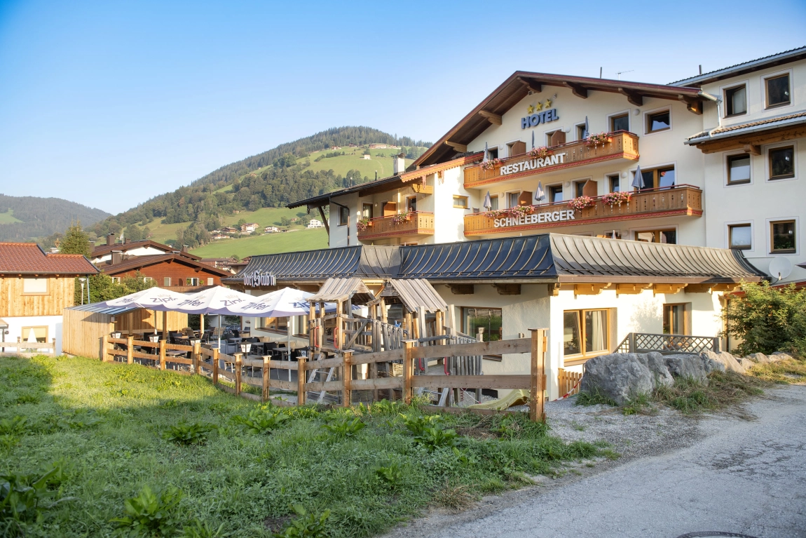 Sitzplatz auf Hotelterrasse mit Blick auf Wiese, Holzzaun, umliegende Gebäude und bewaldete Hügel.