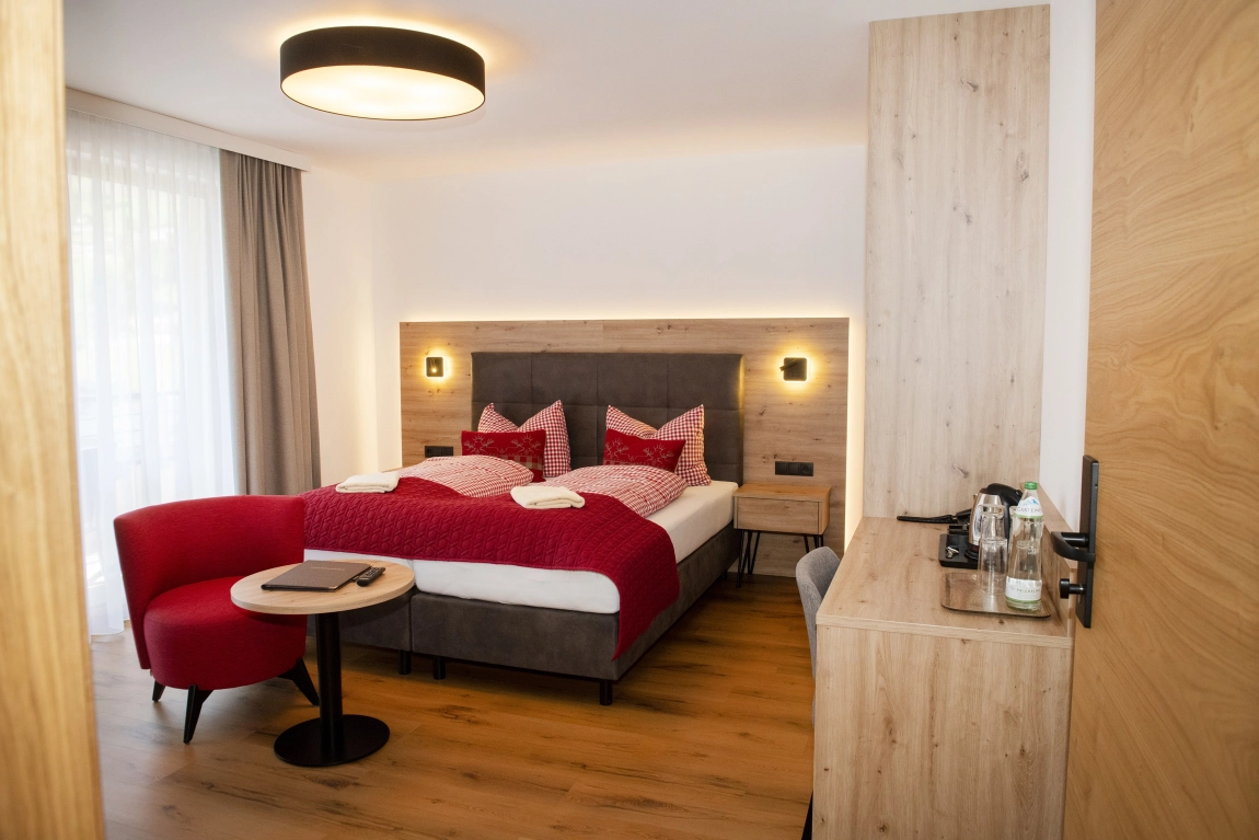 Helles Zimmer mit Doppelbett, roter Sitzecke, Holzschreibtisch mit Wasserkocher, Vorhängen und Deckenleuchte auf Holzfußboden.