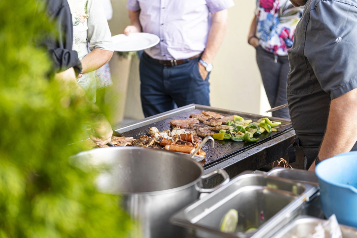 Grillstation im Außenbereich mit Fleisch, Gemüse, Töpfen und wartenden Personen mit Tellern.