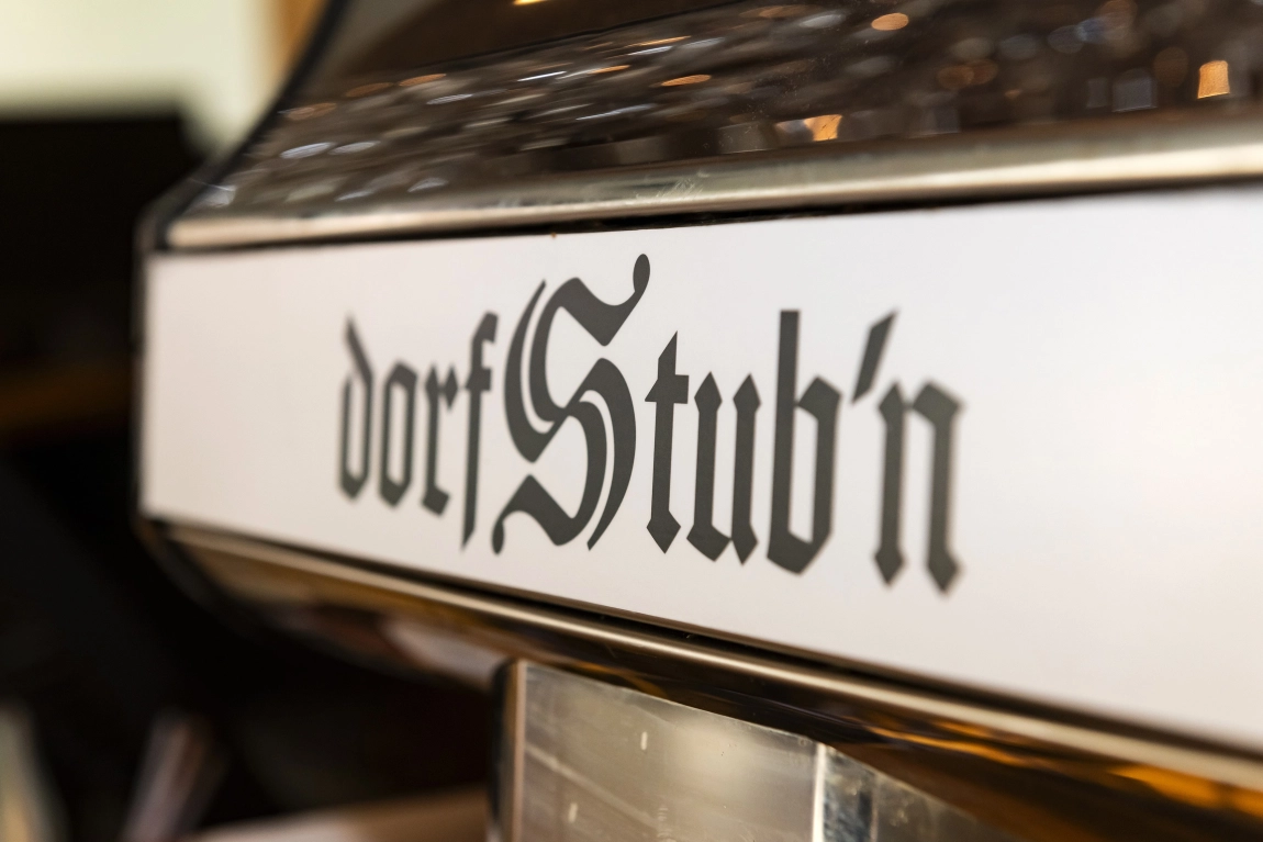„Nahaufnahme des Schriftzugs ‚dorfStub’n‘ in traditioneller Schriftart auf hellem Hintergrund – gemütliches, ländliches Ambiente.