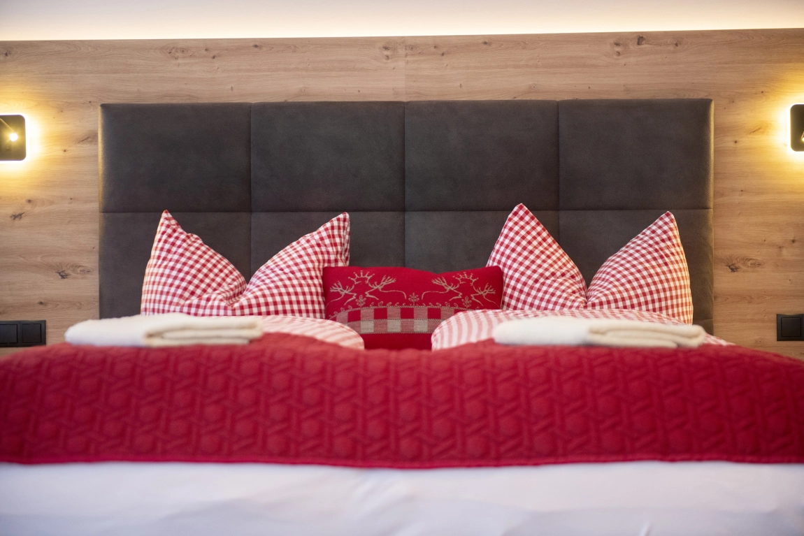 Doppelbett mit rotem Bezug, rot-weiß karierten Kissen und modernem gepolstertem Kopfteil vor Holzrückwand.