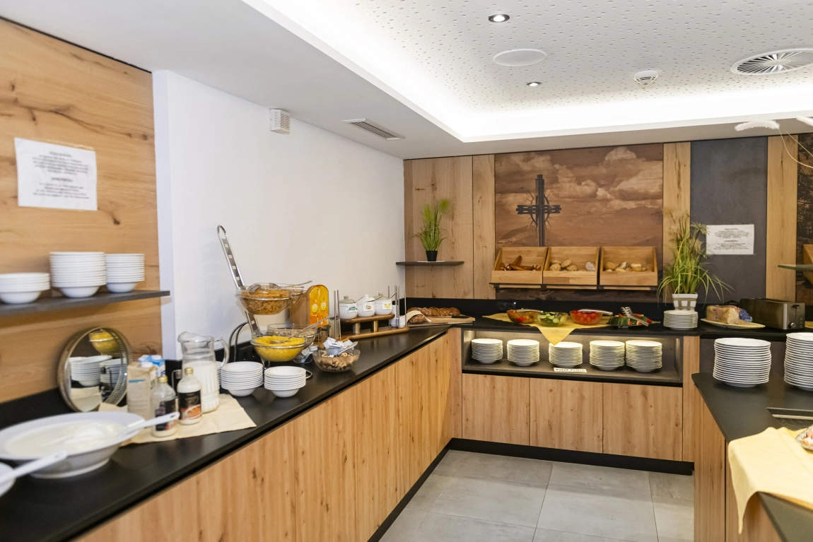Frühstücksbuffet im Hotel mit Brotkörben, Müslibar, Getränken, Tellern, Besteck und Wandbild mit Kreuzmotiv.