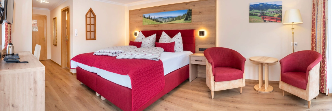 Geräumiges Zimmer mit rotem Doppelbett, zwei Sesseln, Holz­tisch, Bild mit Bergwiese und hellem Holz­boden.