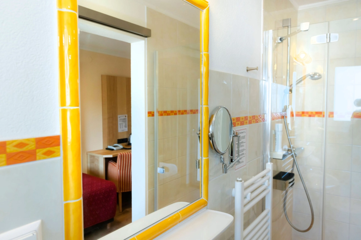 Helles Bad mit Dusche, gelb umrahmtem Spiegel, Handtuchheizung, Duschkopf und Blick ins angrenzende Zimmer.