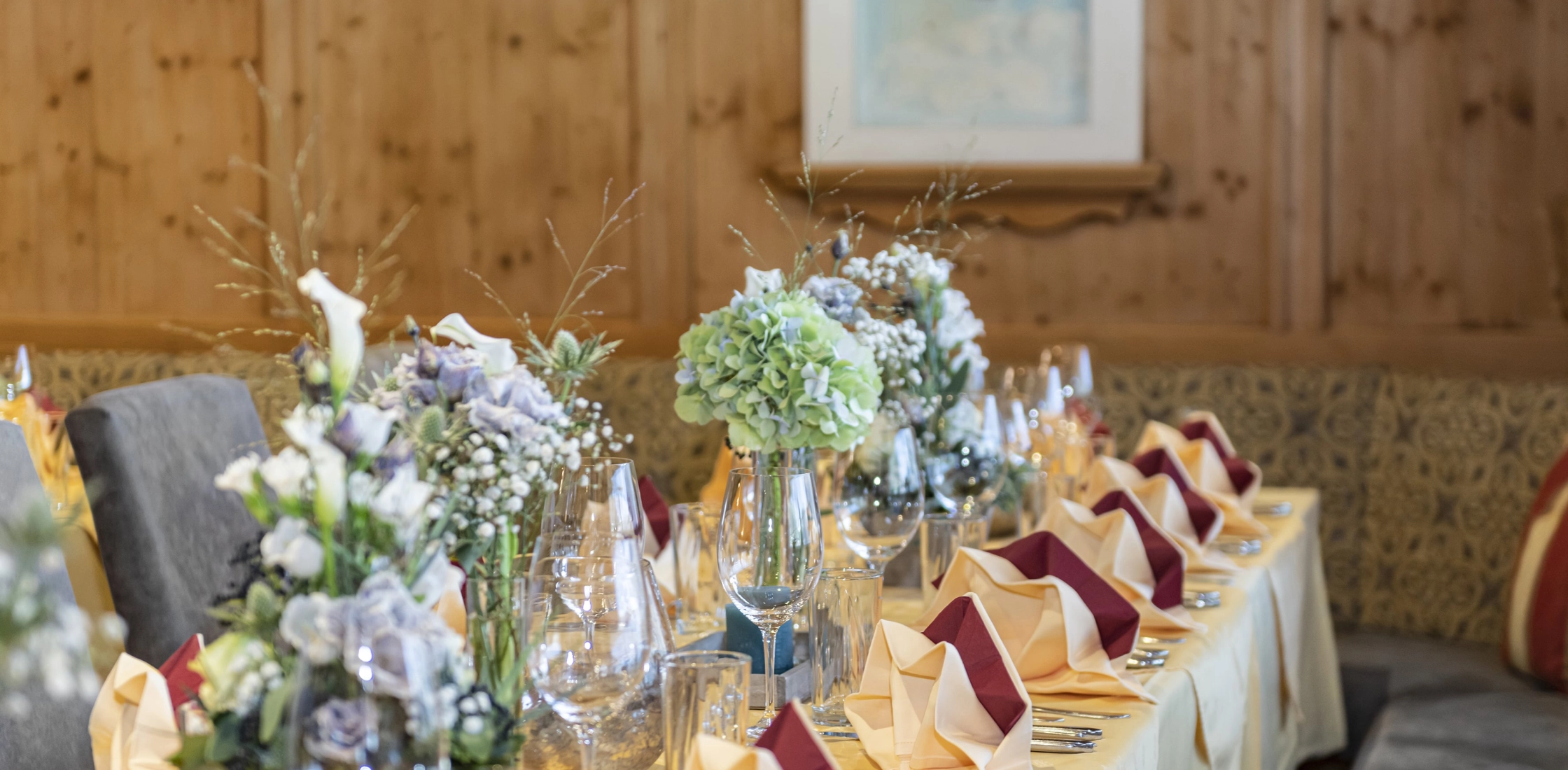 Festlich gedeckter Tisch mit gelber Tischdecke, Blumensträußen, gefalteten Servietten, Gläsern und Holzvertäfelung im Hintergrund.