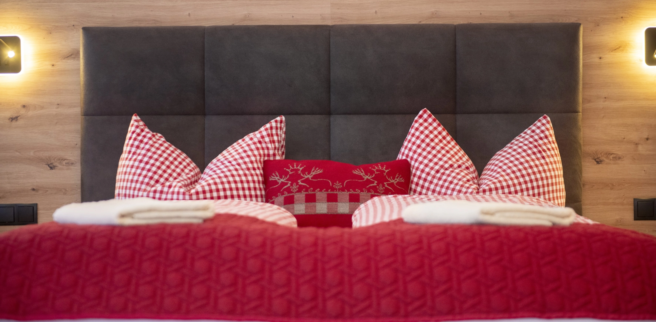 Doppelbett mit gepolstertem Kopfteil, roten Decken, karierten Kissen und gemütlicher Beleuchtung an Holzrückwand.